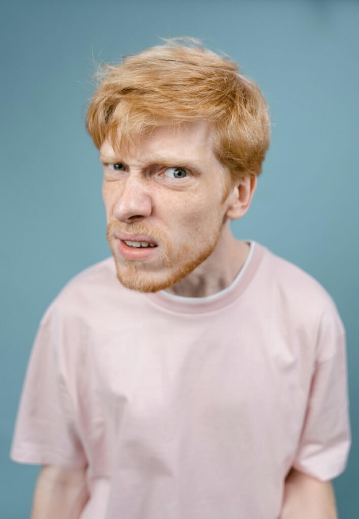 Redhead man with a comical expression wearing a pink shirt against a blue background.